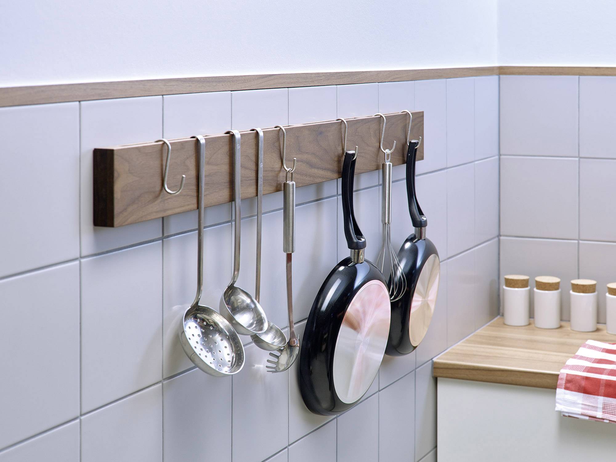 Hanging kitchen utensils on a hook rail: ladle, spatula, two frying pans. Background of white tiles and worktop.