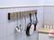 Hanging kitchen utensils on a hook rail: ladle, spatula, two frying pans. Background of white tiles and worktop.