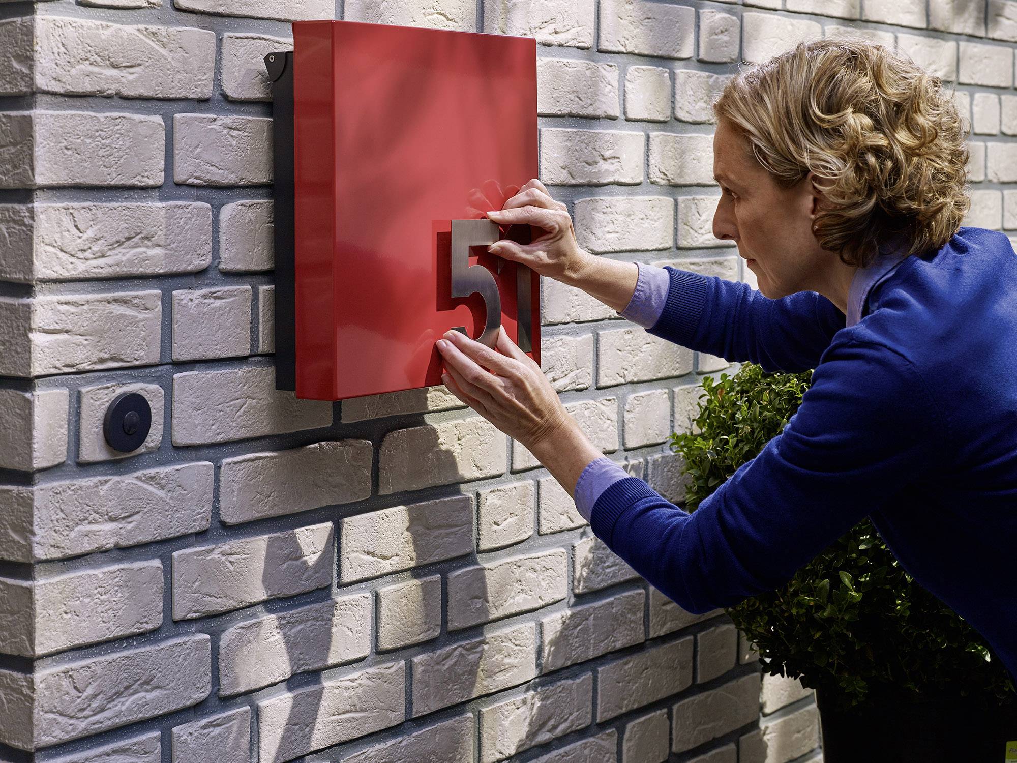 A woman attaches the number '5' to a red sign on a white brick wall beside an entrance door.