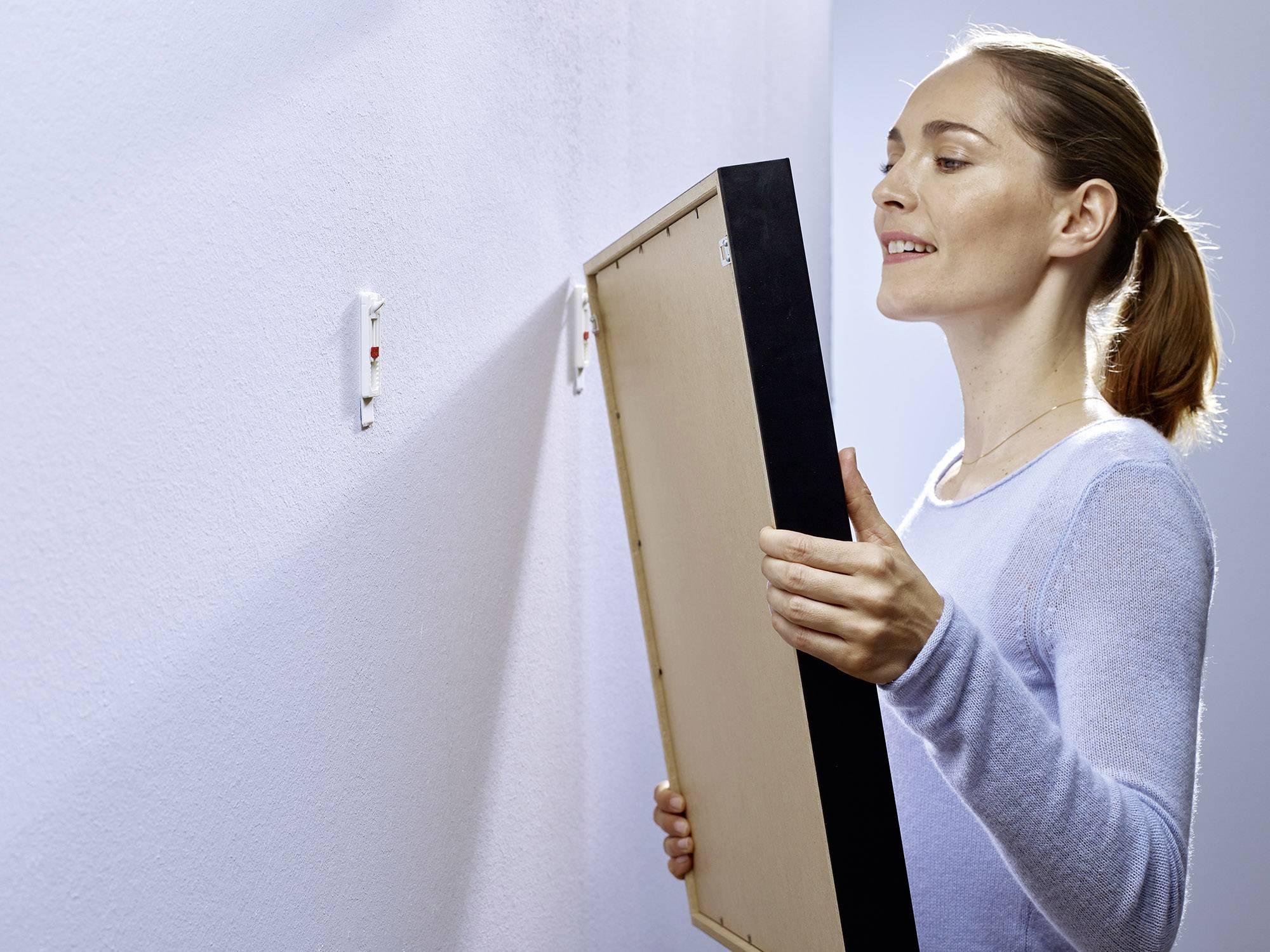 A woman is hanging a picture on a wall. She holds the frame with both hands while aligning the picture to the already installed hooks.