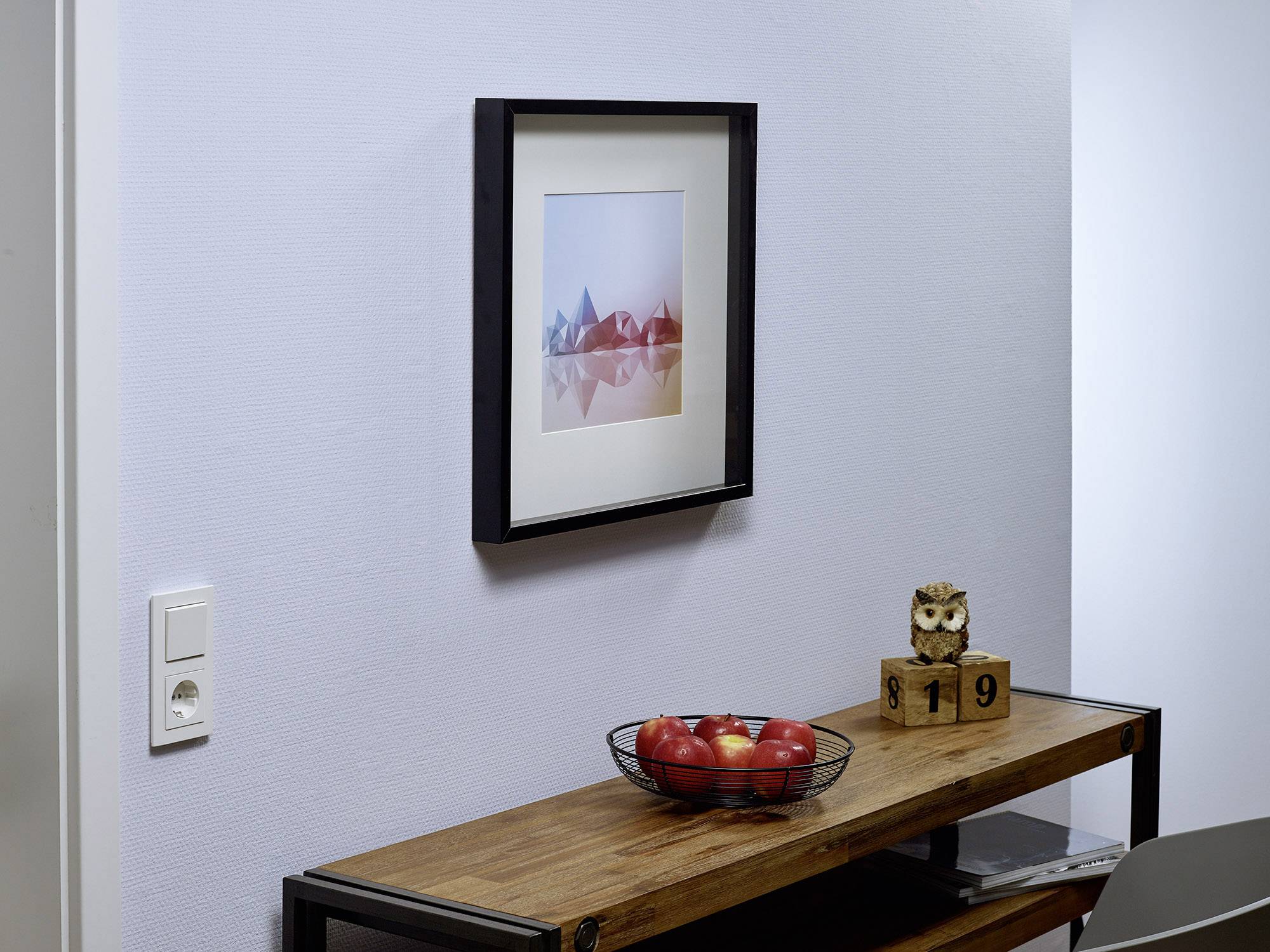 A framed picture on the wall above a wooden table with a basket full of red apples and a cube-shaped calendar.