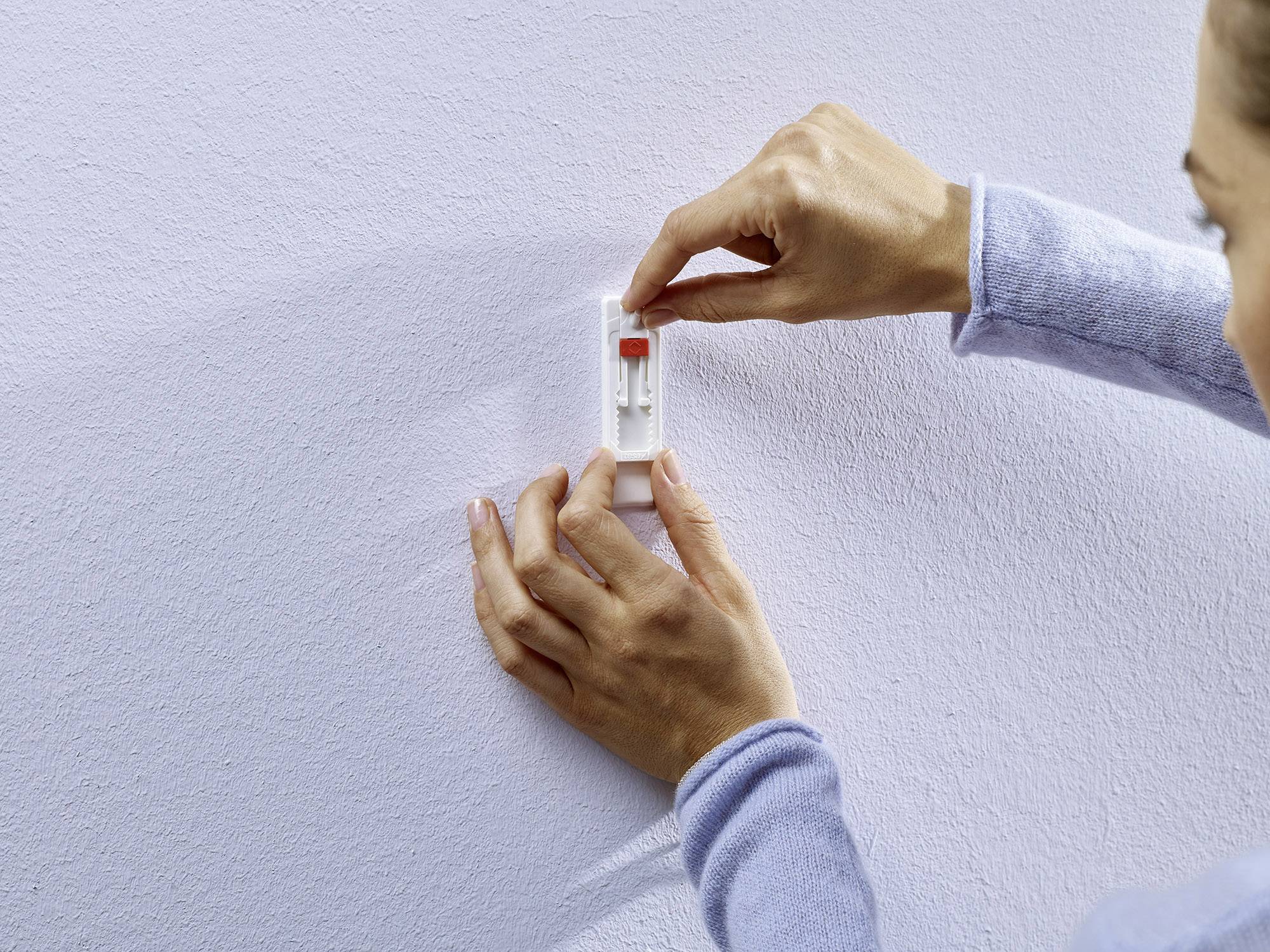 A person is attaching a self-adhesive hook to a white wall.