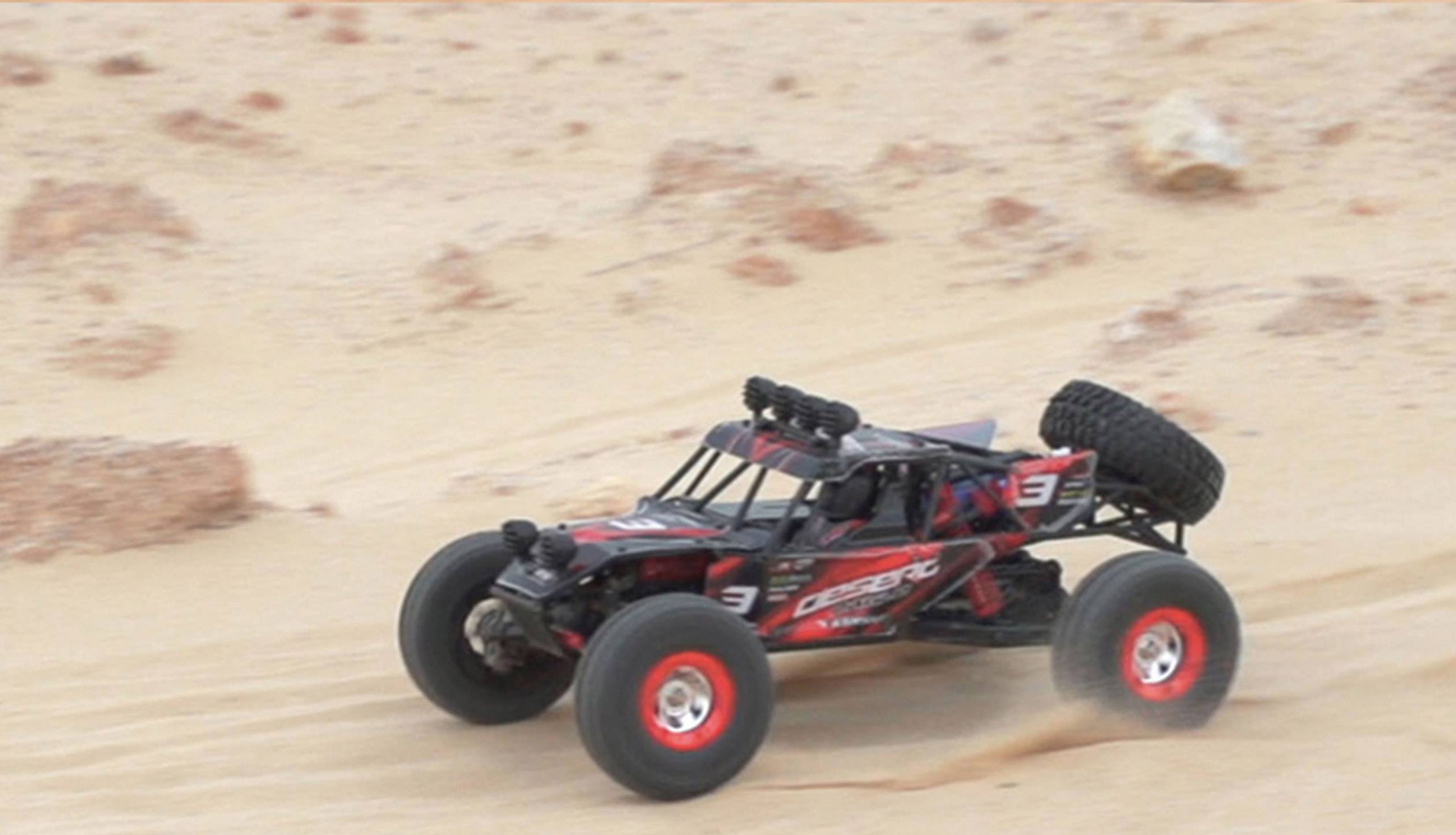 A remote-controlled car speeds across a sandy desert landscape. Large tyres kick up dust.
