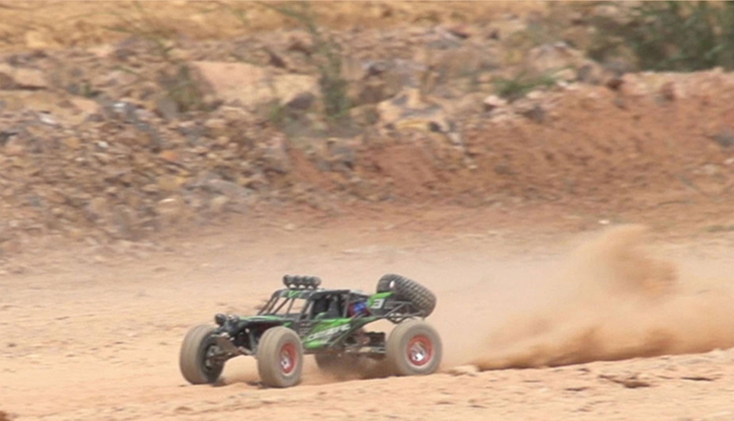 A remote-controlled car speeds across a sandy, uneven surface, kicking up dust. Background with rocks and earth.
