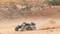 A remote-controlled car speeds across a sandy, uneven surface, kicking up dust. Background with rocks and earth.