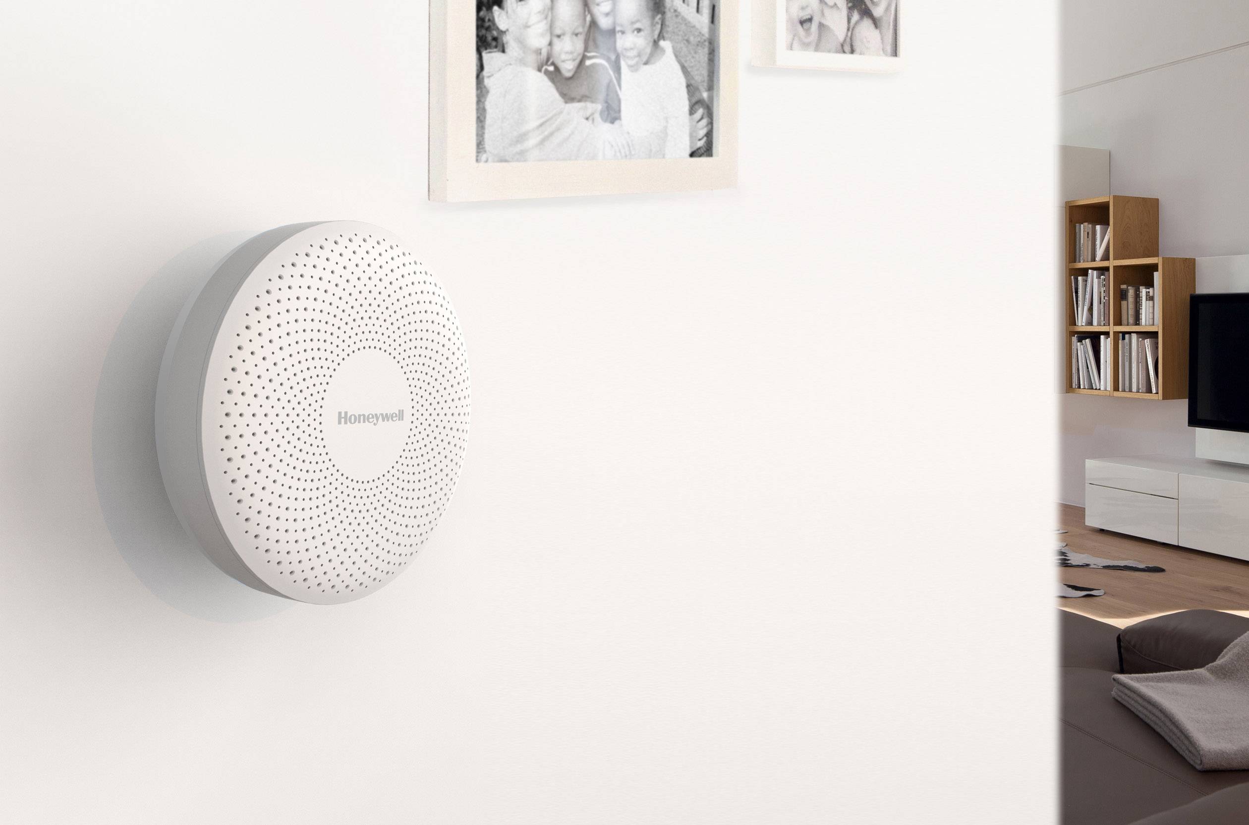 A round, white smoke detector is mounted on a wall in a living room. In the background are bookshelves and a television.