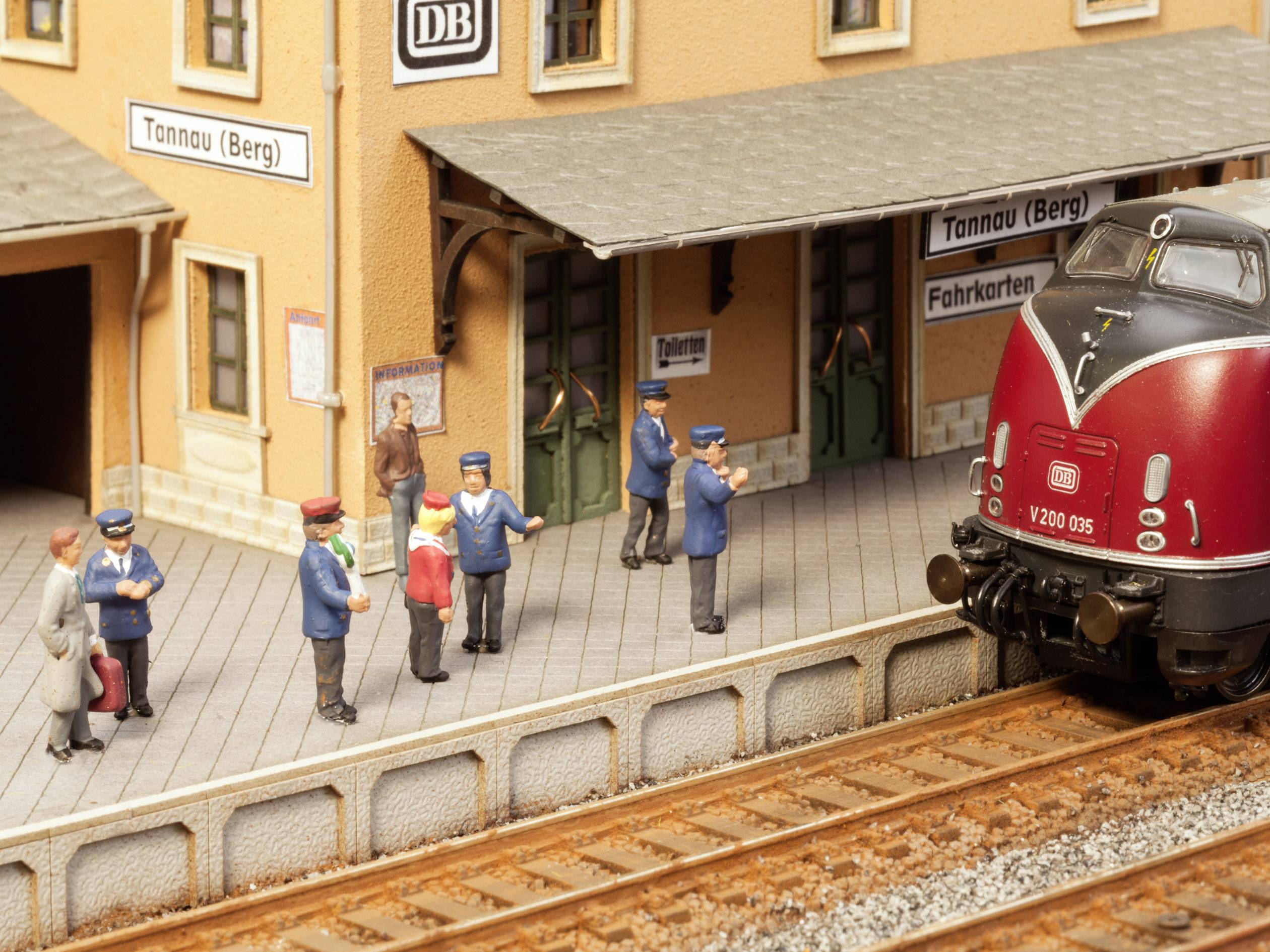 A miniature railway scene depicts a red locomotive on a track in front of a station with figures of passengers and railway staff.