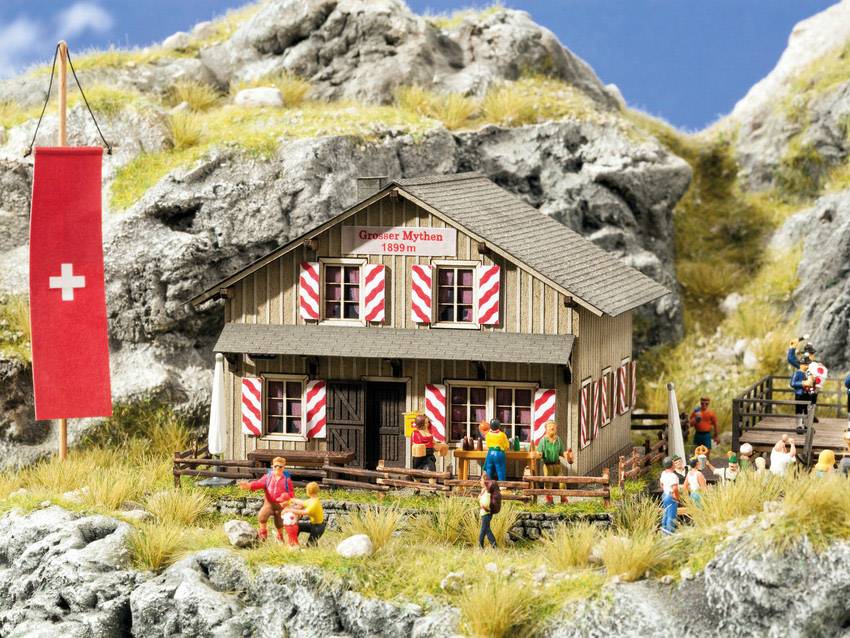 Alpine miniature model of a building with red and white shutters, people, and a Swiss flag in a mountainous landscape.