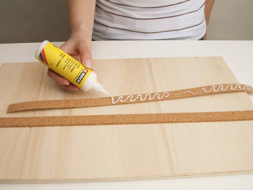 A person is applying adhesive to a cork strip lying on a wooden board. The cork is being brushed with an undulating line.