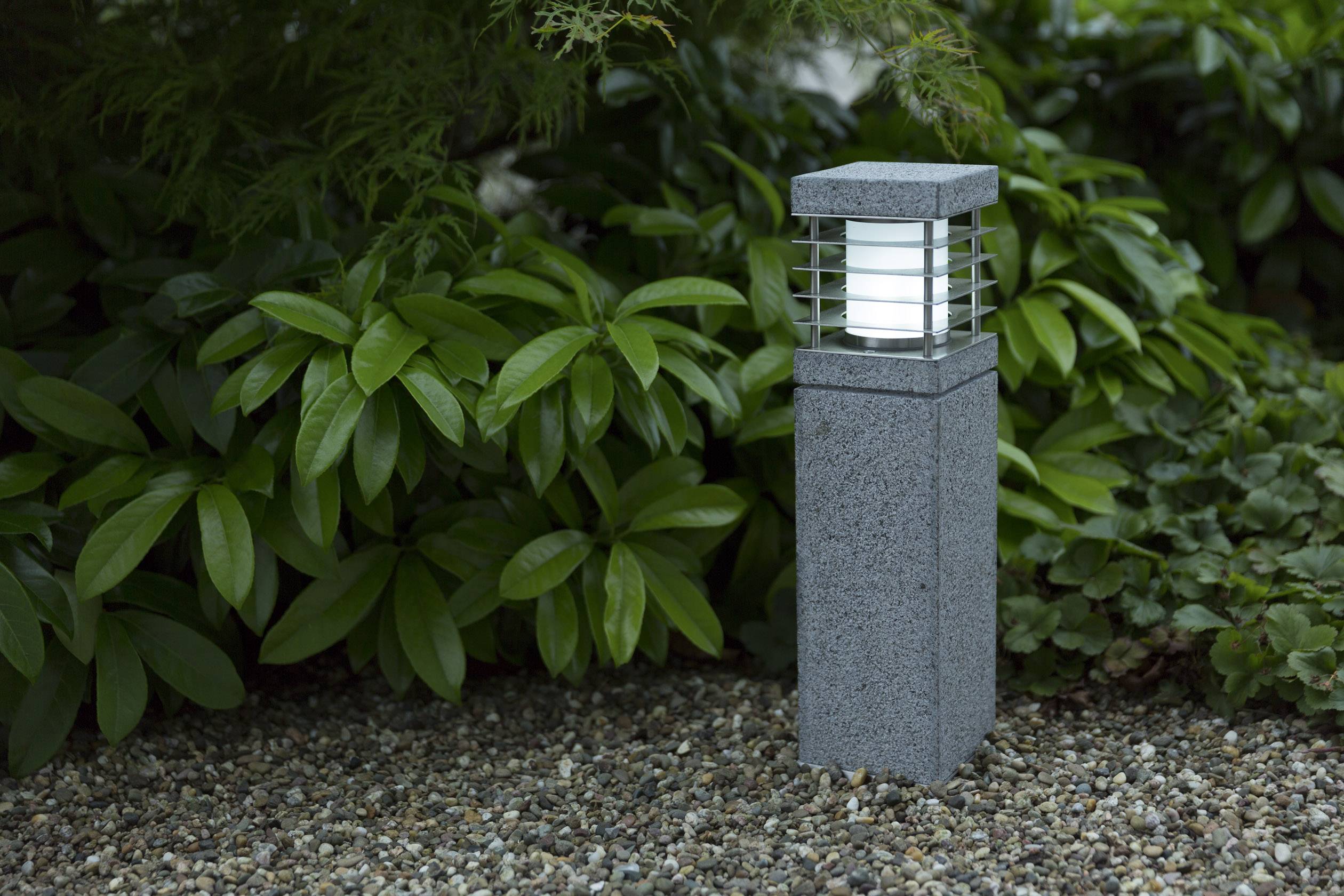 A stone garden lantern stands on a gravel path, surrounded by green leaves, and glows softly in the twilight.