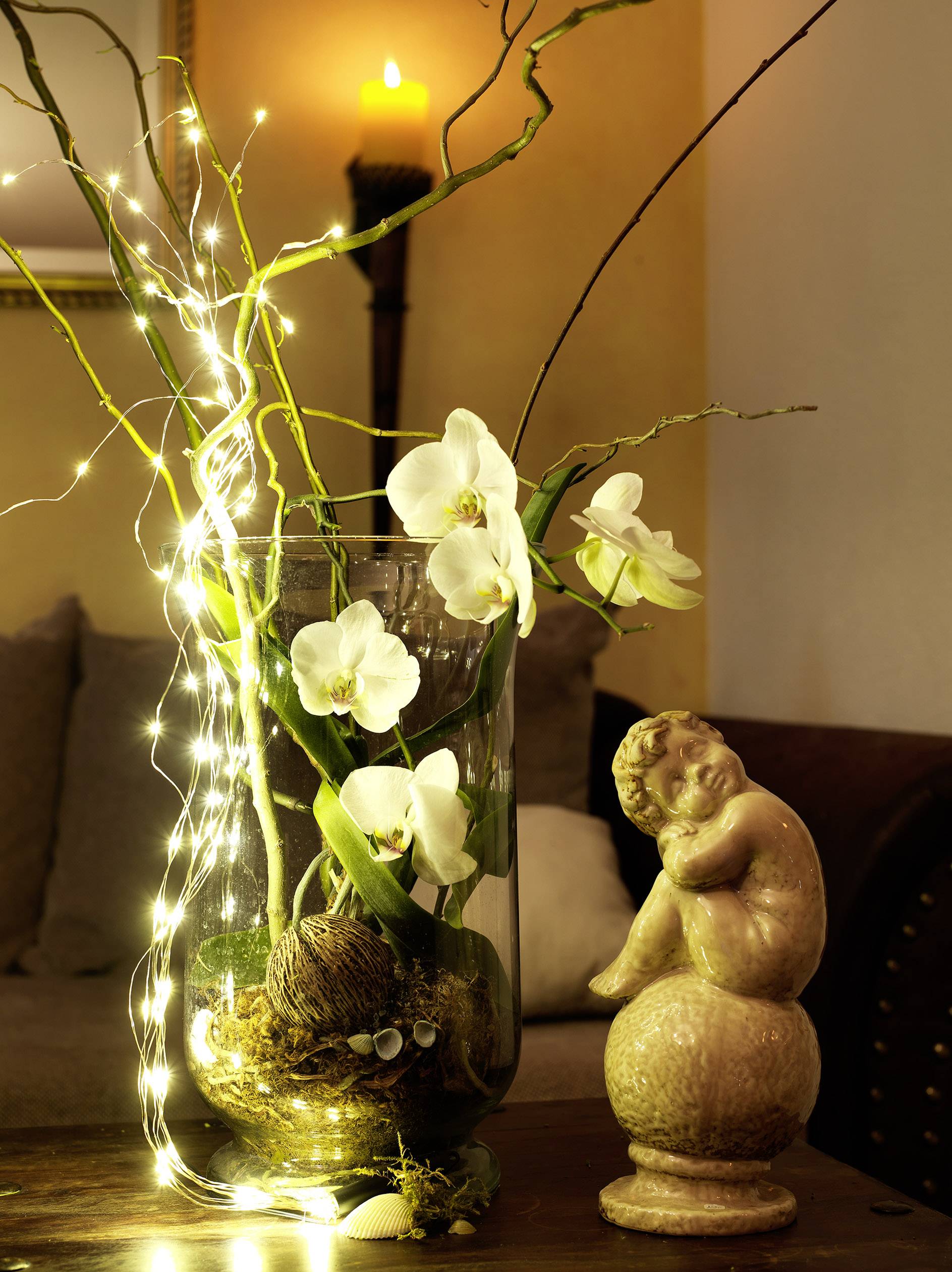 A vase with white orchids and fairy lights sits on a table. Beside it, an angel statue is positioned. In the background, a candle is burning.
