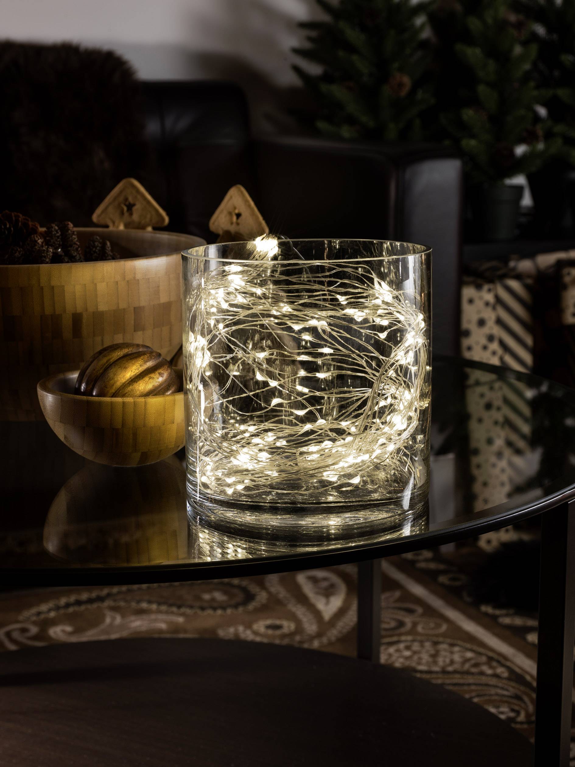 A glass bowl on a table, filled with twinkling fairy lights. Beside it sits a wooden bowl with decorative baubles.