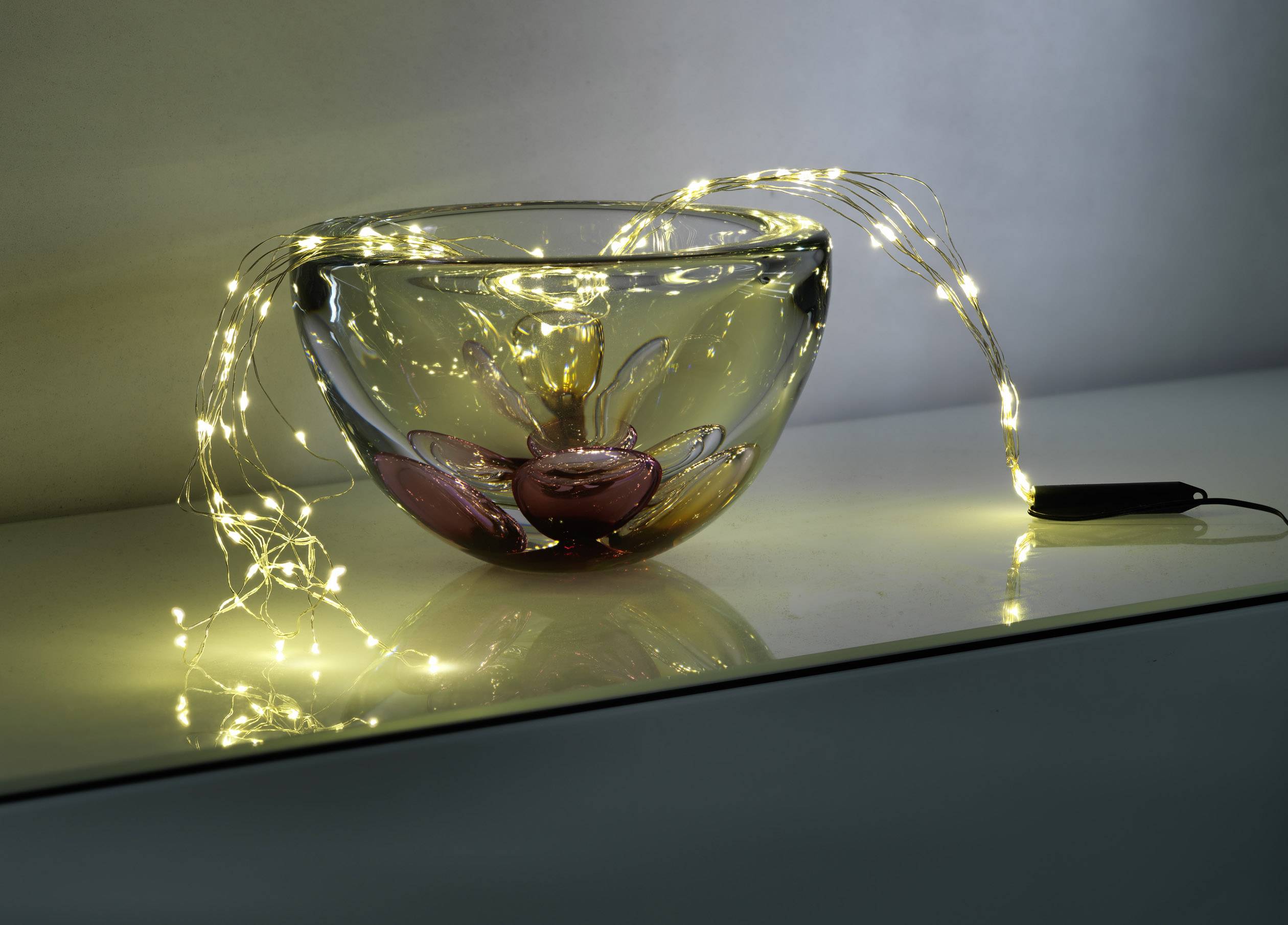 A glass bowl with colourful accents, decorated with twinkling fairy lights, sits on a smooth, white surface.