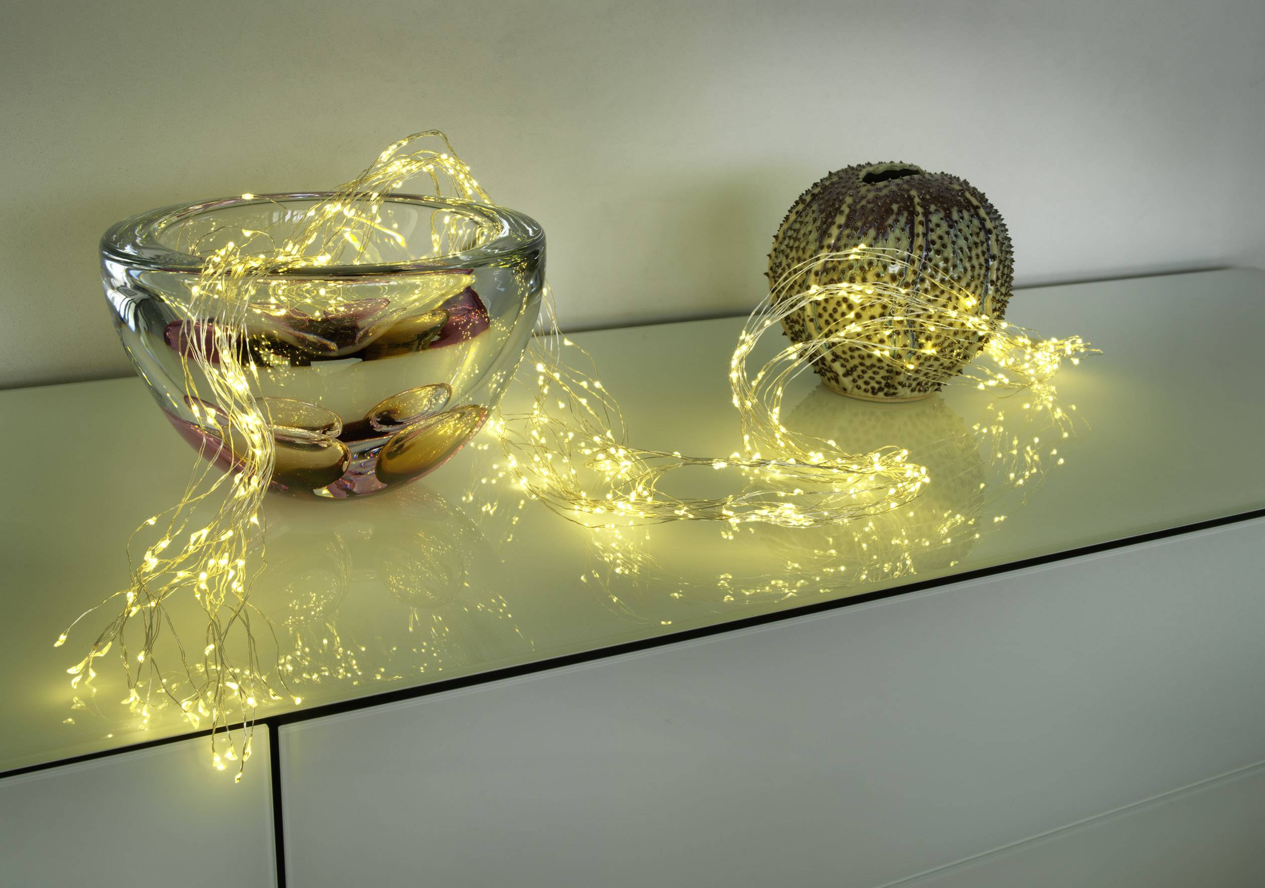 On a white table stands a bowl with glass decorations and beside it a glowing string of lights in a warm yellow tone.