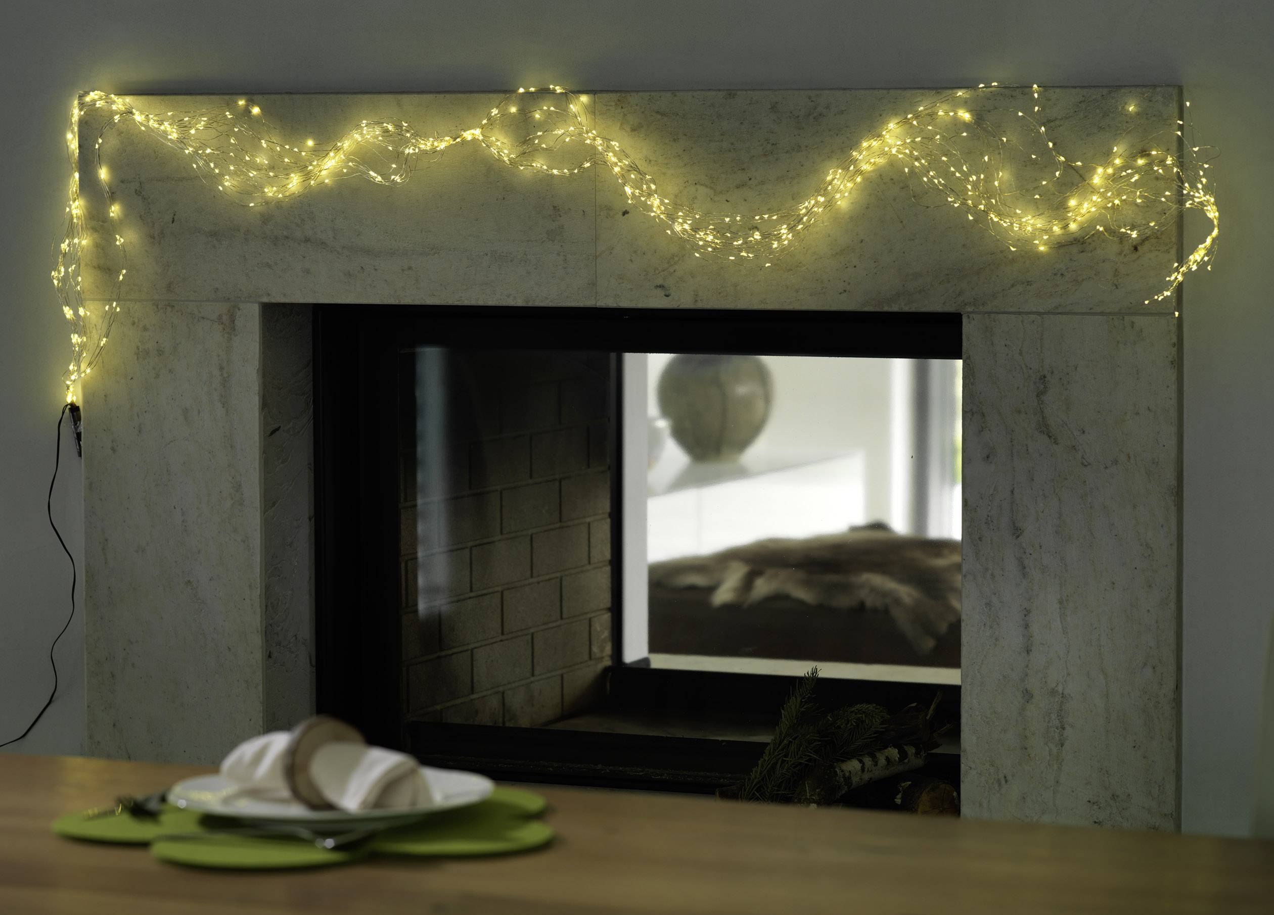A fireplace with a white marble surround is decorated with a glowing yellow string of lights. In the foreground is a laid table.