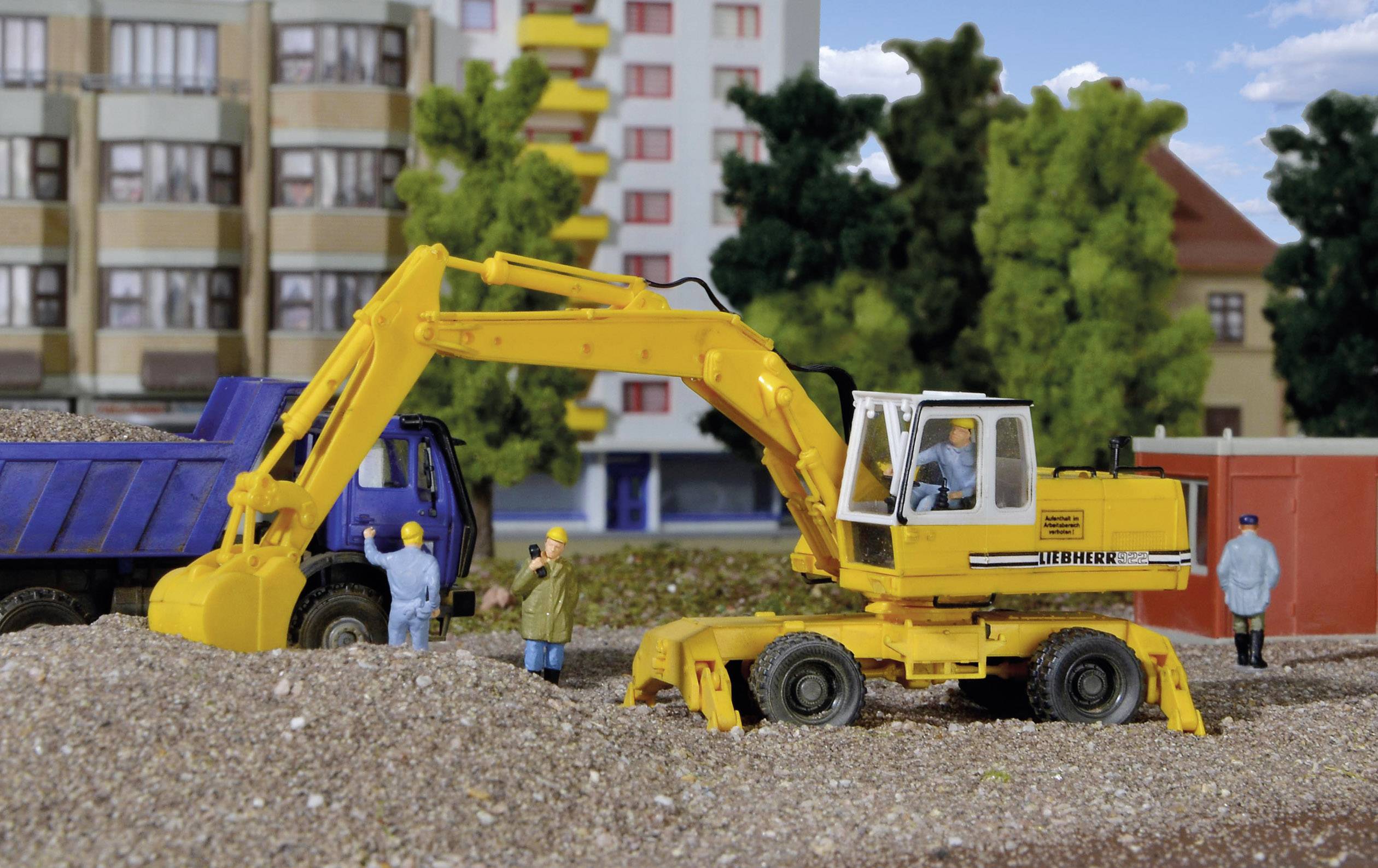 A yellow excavator is lifting gravel into a blue lorry. Three construction workers are standing nearby. In the background, residential houses and trees are visible.