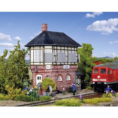 A red model railway building with half-timbered style and human figures in the foreground. A red train model is also visible.