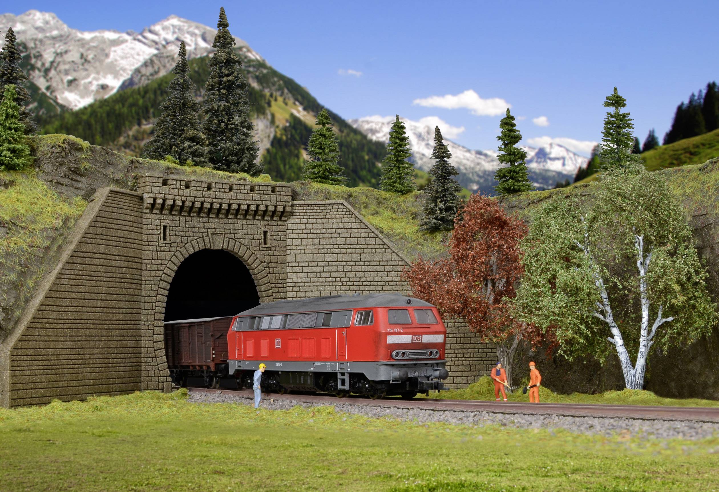 A red locomotive emerges from a tunnel in a picturesque mountain landscape with trees and two people wearing orange clothing.