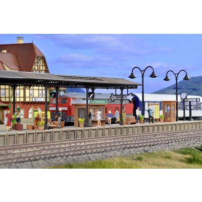 A miniature railway station with a red train on the track. Passengers are waiting on the platform. A timetable and signs are visible.