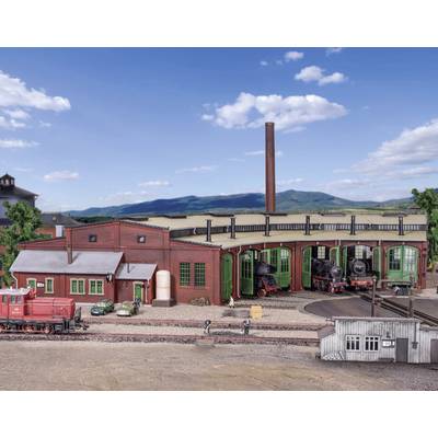 Model railway shows a railway depot with a locomotive shed, two steam locomotives and a red diesel locomotive in the foreground against a hilly backdrop.