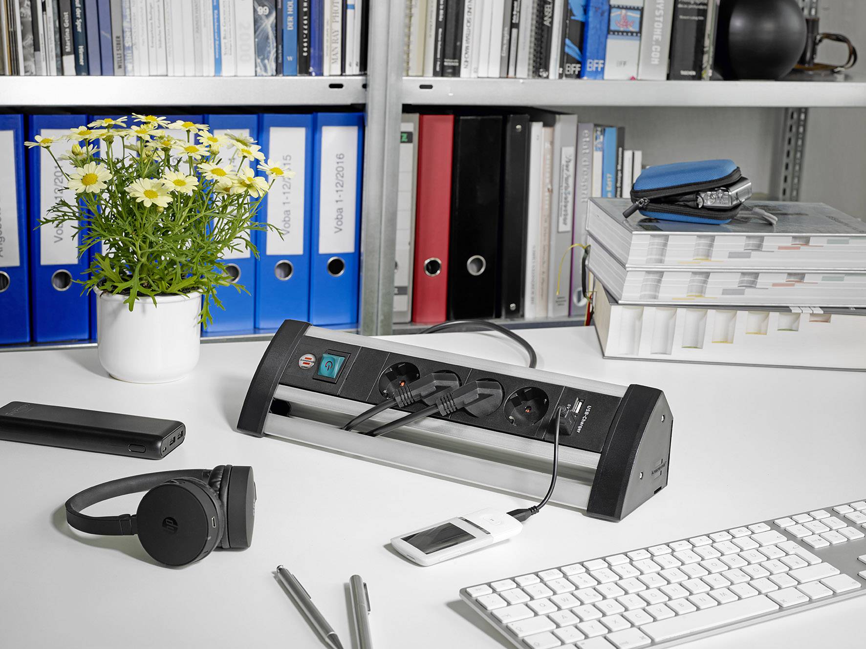 A desk with books, folders, a power strip, headphones, a smartphone, a keyboard and a bouquet of flowers.