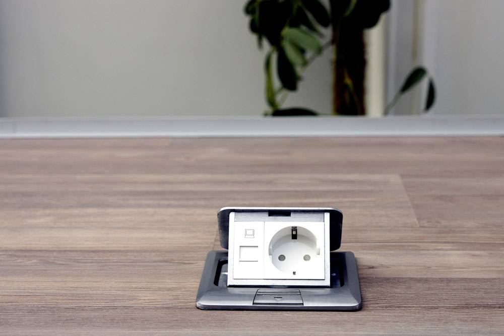 A wooden floor reveals an open electrical socket; a plant is blurrily visible in the background.