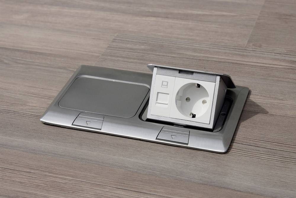A floor socket with an open hinged lid is located in a wooden floor. The socket and connections are visible.