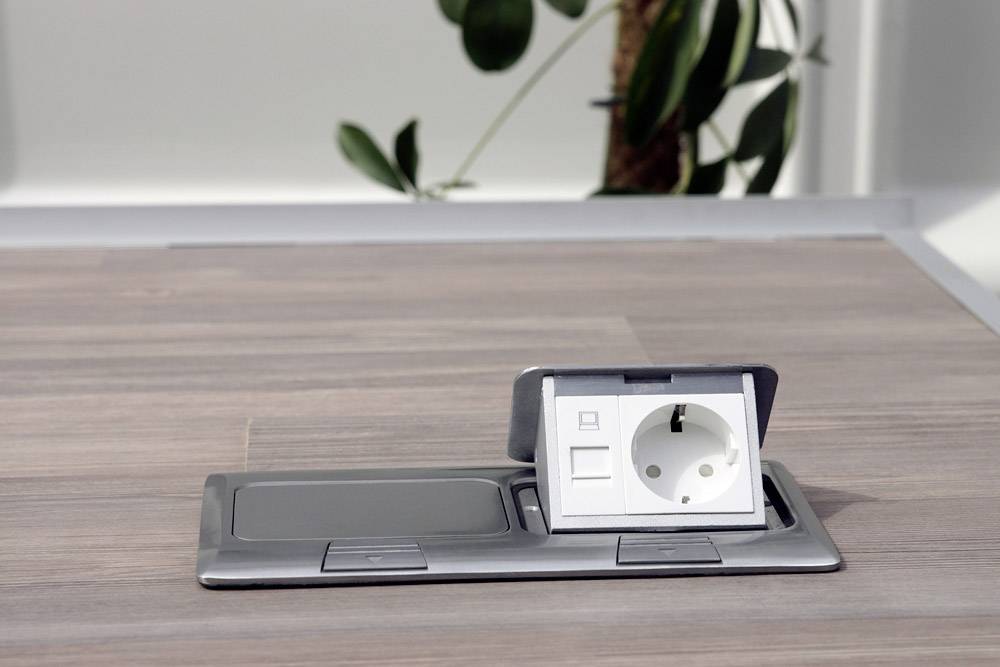 Socket embedded in table surface, surrounded by wood grain pattern. A plant is slightly out of focus in the background.