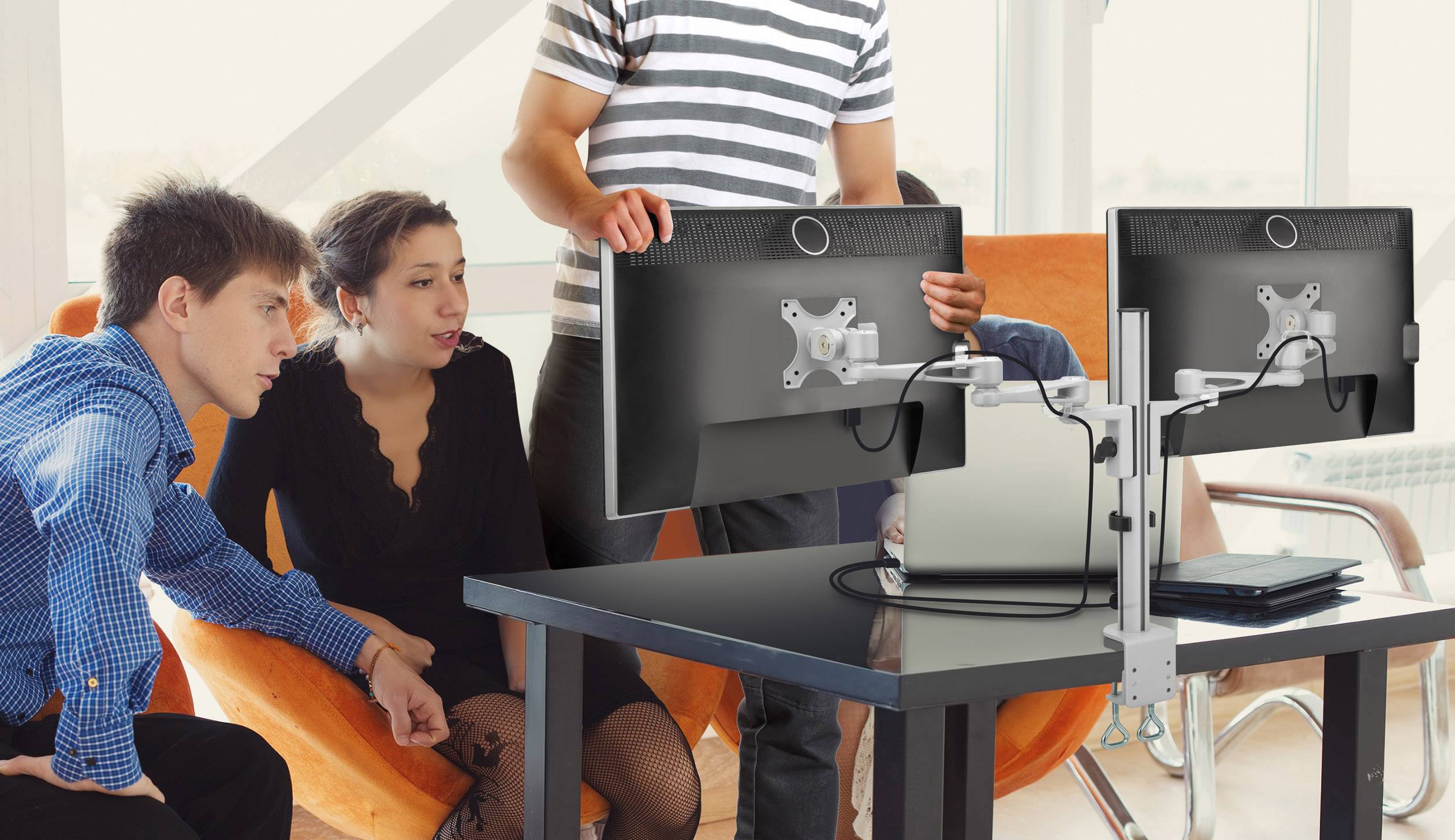 Four people are sitting around a table with two monitors. One person is standing and pointing at a screen. They appear to be discussing something.