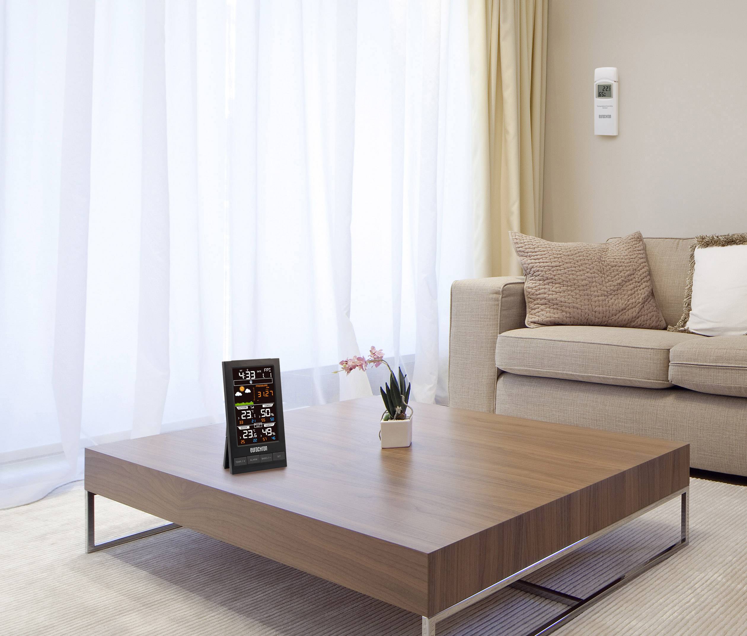 A living room with a wooden table, on which a digital weather station sits. A plant is positioned beside it. A sofa is in the background.