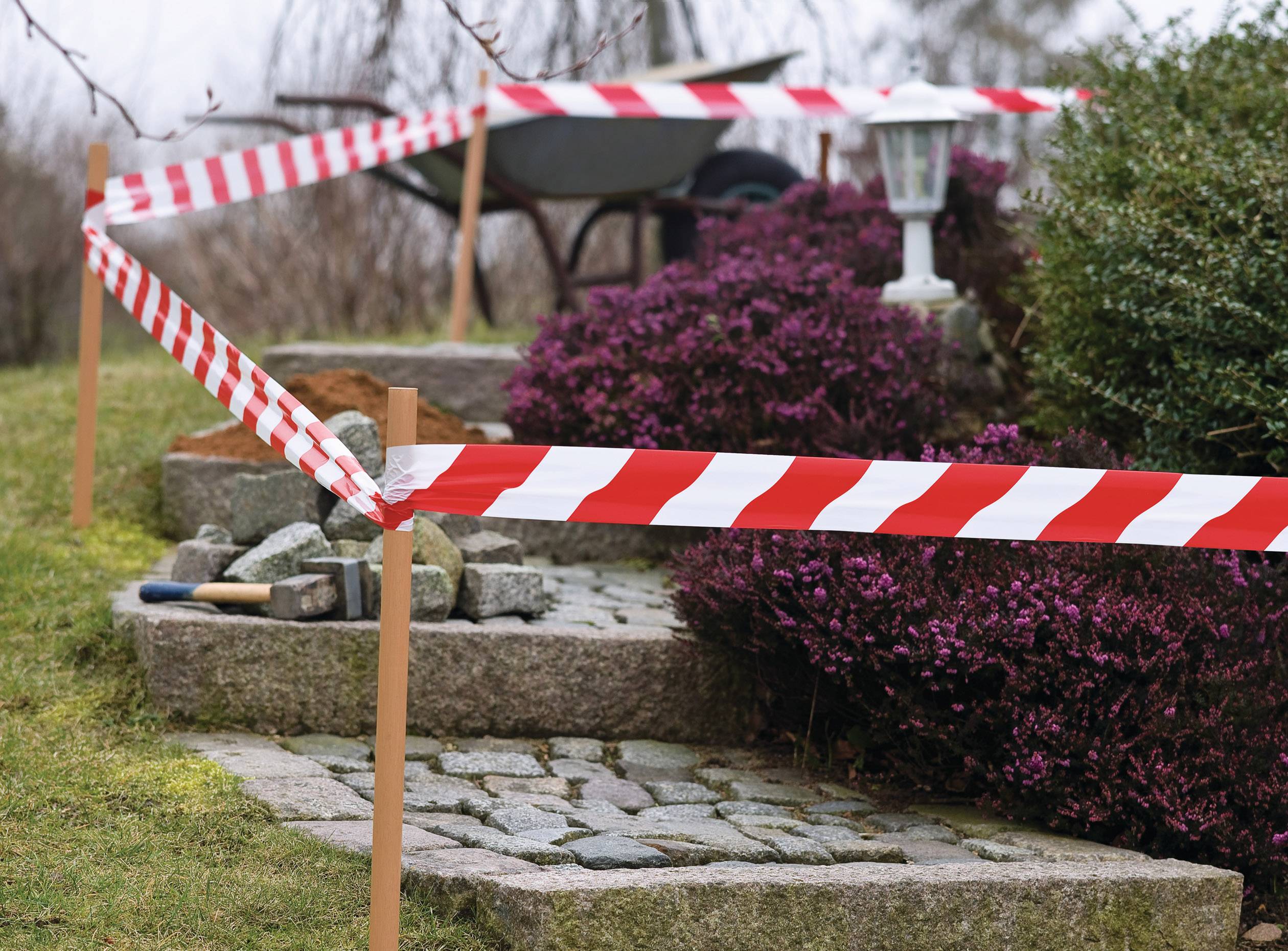 Barrier tape surrounds a garden path marked with red and white stripes. A wheelbarrow, several plants, and a lantern can be seen.