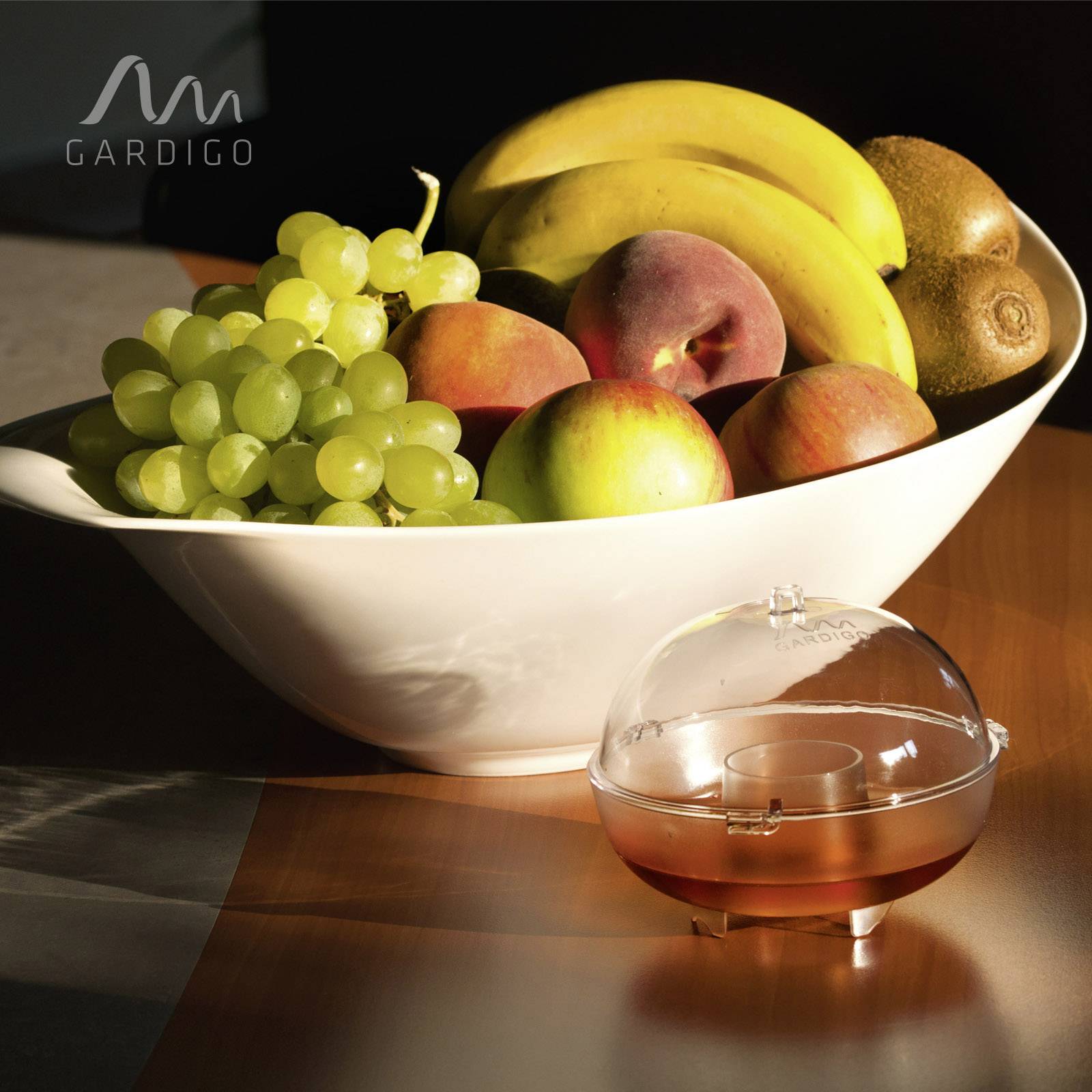A fruit bowl with bananas, apples, and grapes sits on a table. Next to it is a transparent, round container with unknown contents.