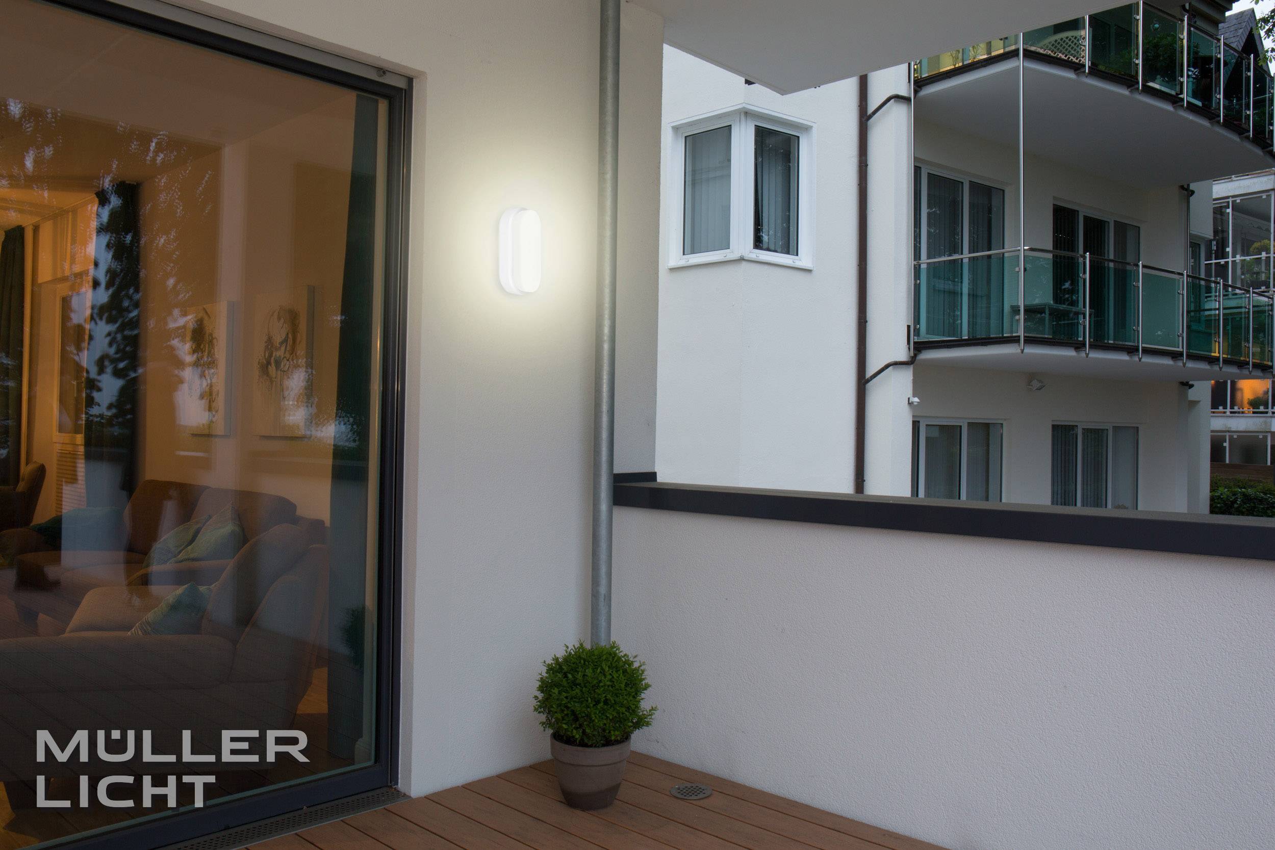 A illuminated balcony at night with a bright light on the wall. In the background, a modern apartment block with balconies is visible.