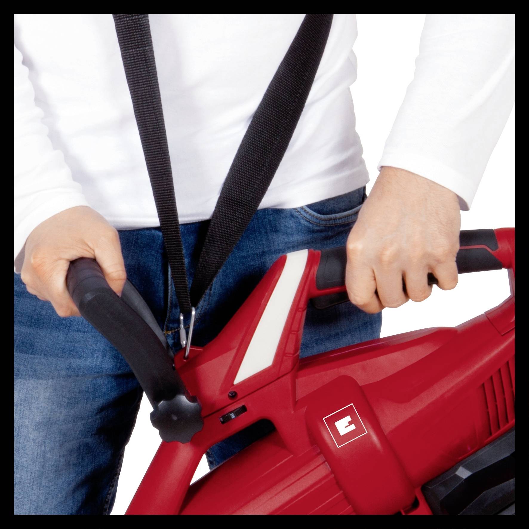 A person is holding a red leaf blower with both hands, ready for use. A black carrying strap is draped around their neck.