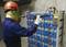 A person in protective clothing is testing the voltage on a series of blue batteries in a concrete room.