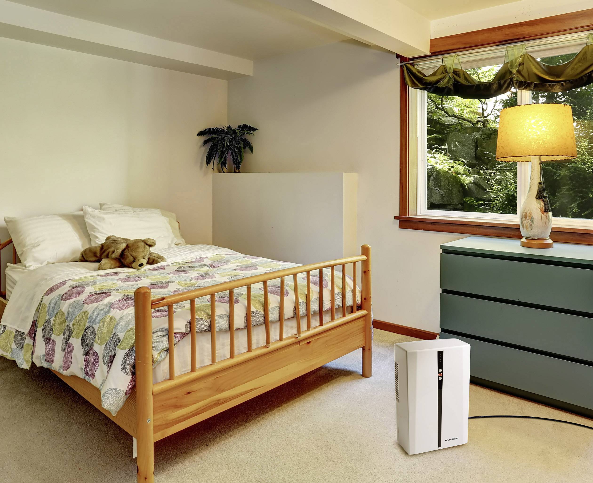 A bedroom with a wooden bed, colourful duvet and a teddy bear on top. To the right stands a chest of drawers with a lamp on it. In the background, a window offers a view of greenery.