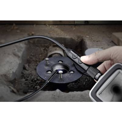 A hand is holding an inspection camera over a dark hole in a concrete floor. The camera is attached to a flexible cable for examining hard-to-reach areas.