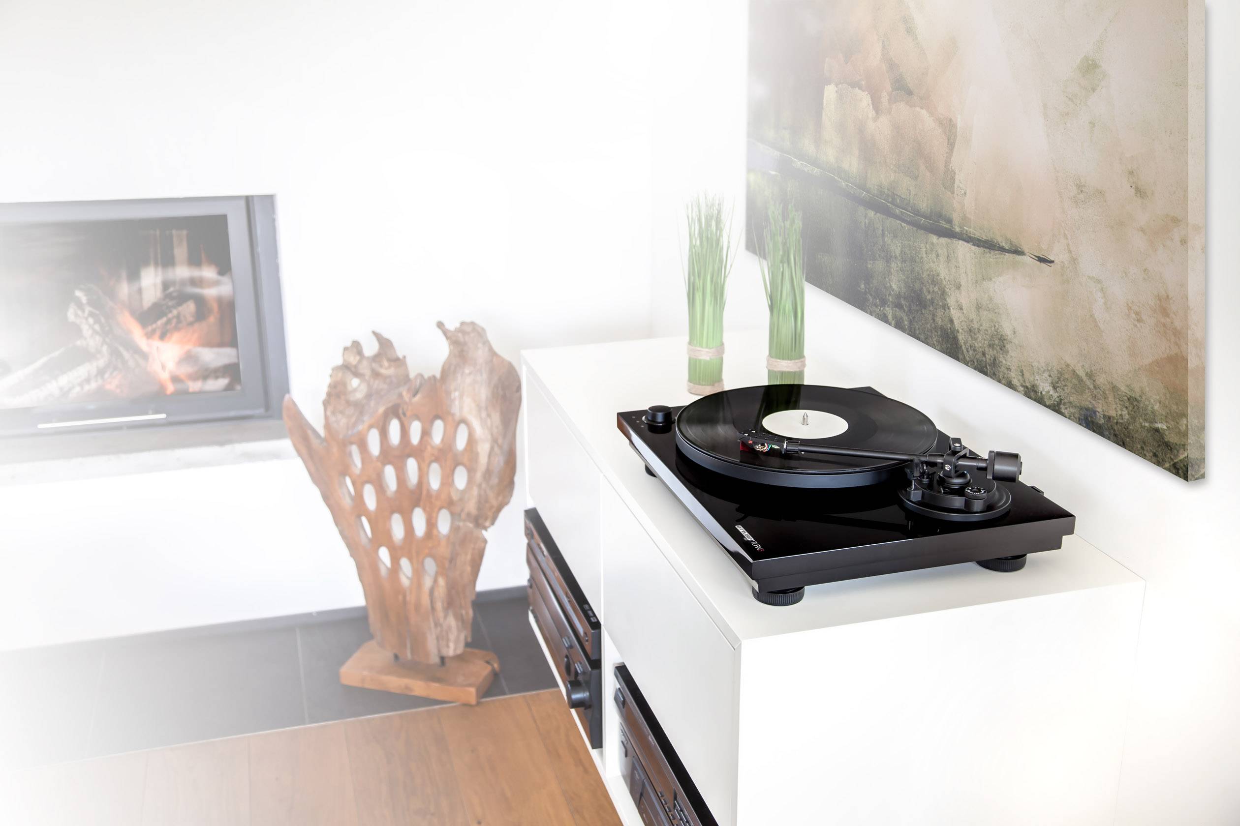 A modern record player on a white cabinet, with green decorative plants beside it. In the background, a fireplace and abstract art.