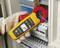 A person is measuring the electrical cable in a control cabinet using a yellow multimeter. The device is showing '247V'.
