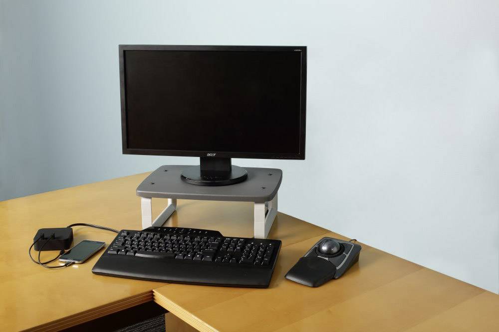 'A computer workstation with a monitor on a stand, keyboard, trackball and smartphone, connected by a charging cable.'