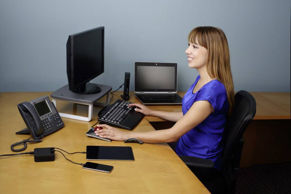 A woman in a blue blouse is sitting at a desk with computers, a telephone, and mobile devices, working with concentration.
