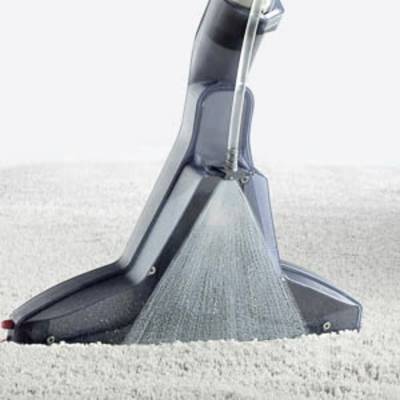 A vacuum cleaner is cleaning a light-coloured carpet. A clear dirty water container shows progress in extracting dirt and water.