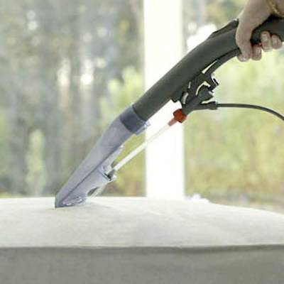 A person is cleaning an upholstery cushion with a vacuum cleaner. The device has a transparent nozzle that sprays and sucks up water.