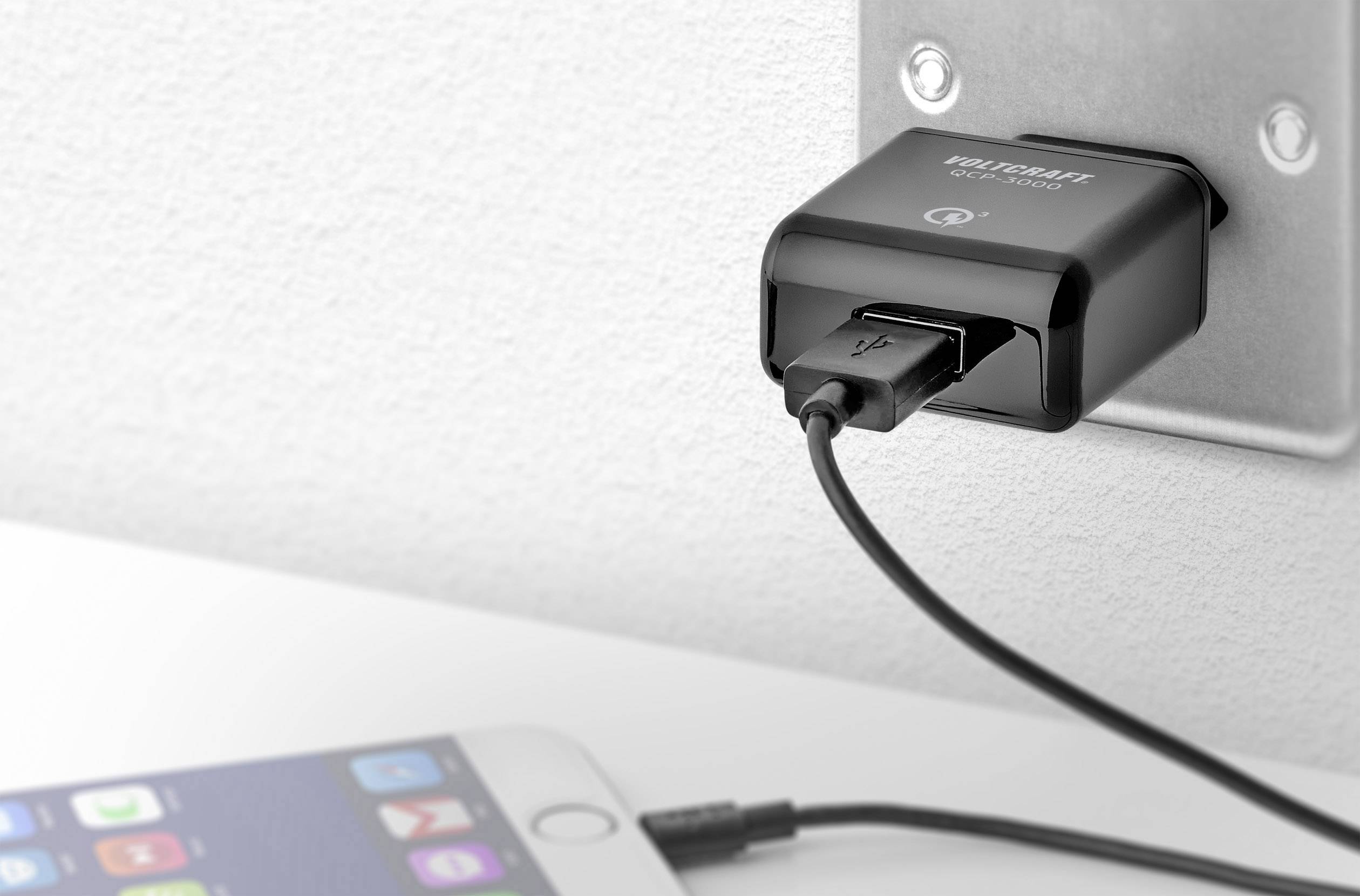 A black USB charger is plugged into a wall socket and is charging a smartphone lying on a white surface.