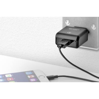 A black USB charger is plugged into a wall socket and is charging a smartphone lying on a white surface.