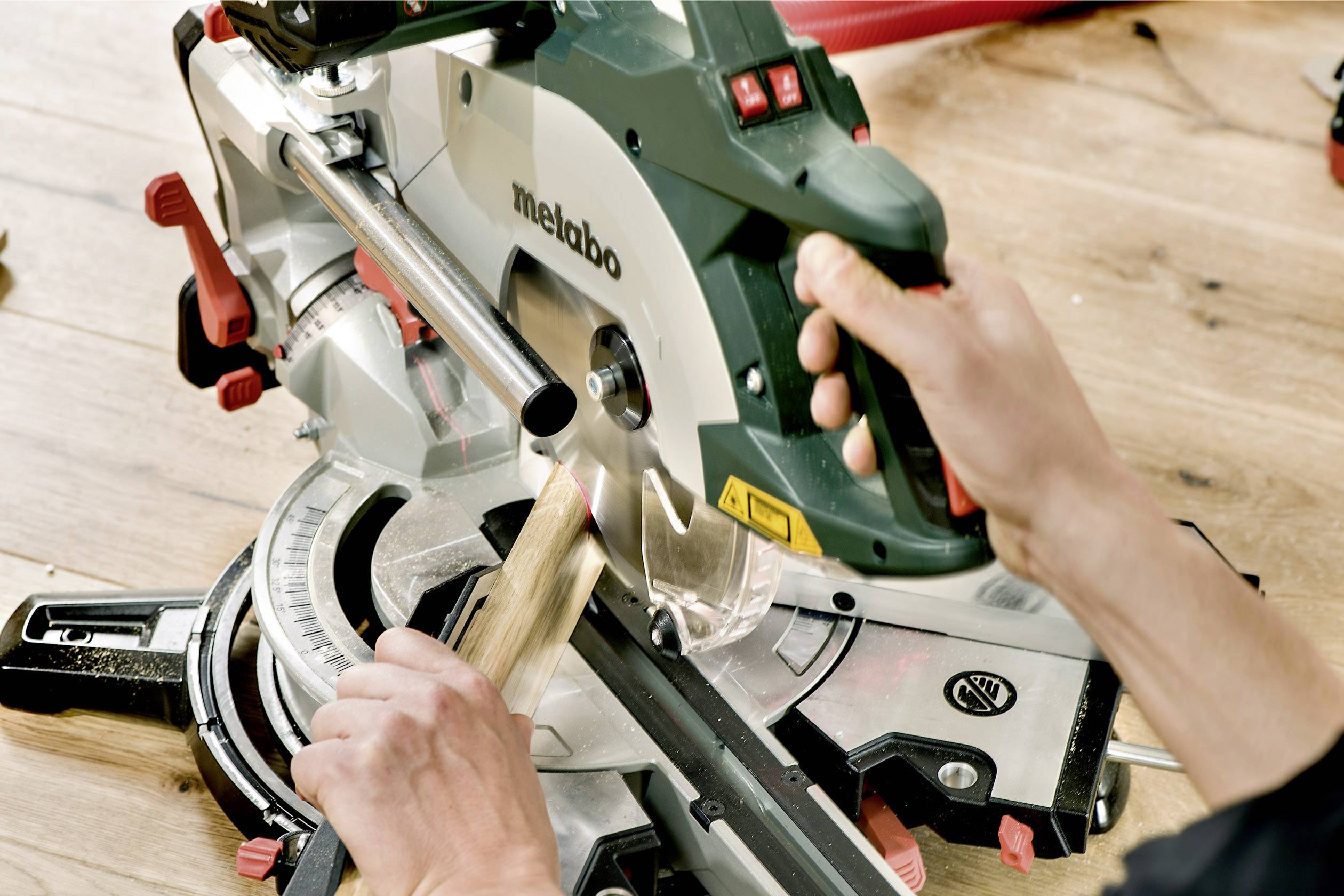 Two hands operate a mitre saw, cutting a piece of wood. The saw is mounted on a wooden workbench.