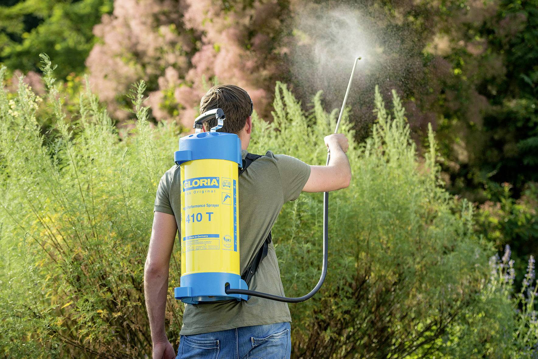 A person is spraying plants with a handheld device in the garden. They are wearing a blue and yellow backpack sprayer.
