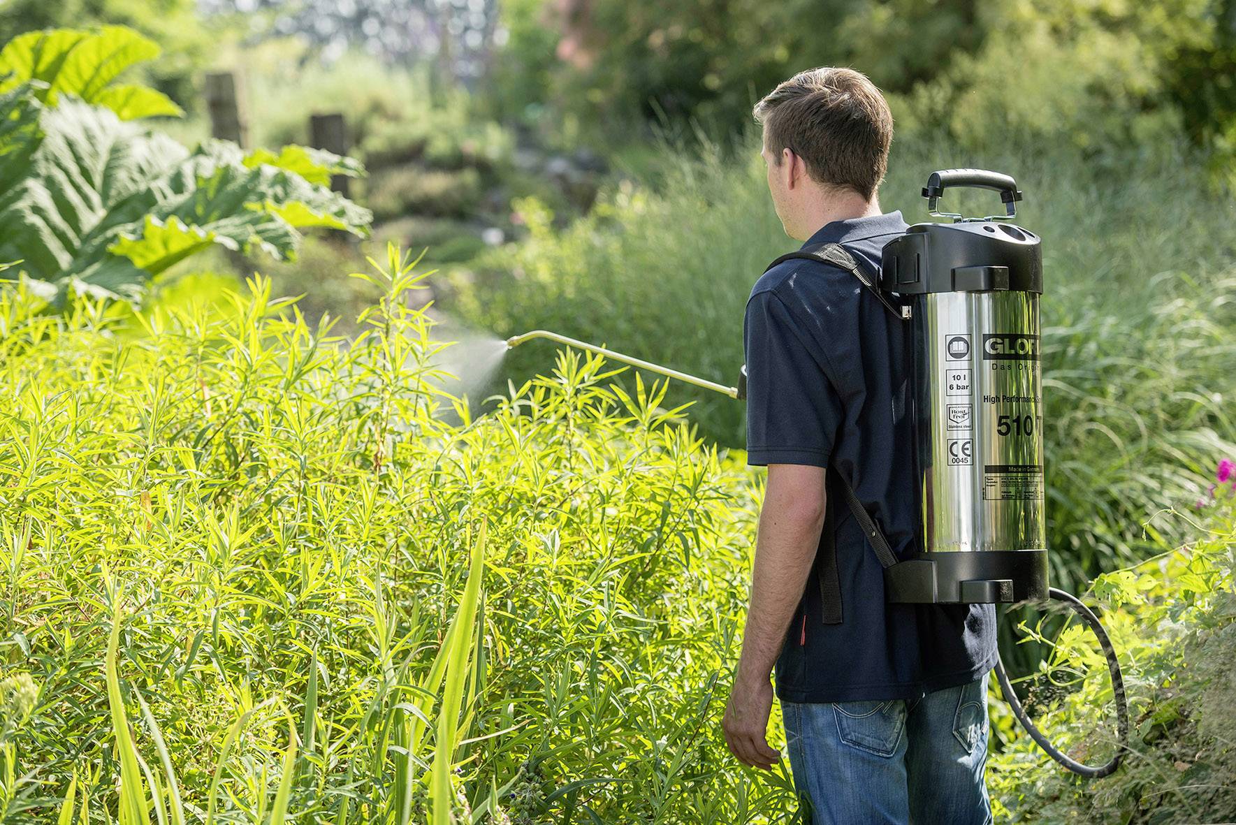 A man with a backpack sprayer is watering plants in a garden. Lush grasses and trees surround him.