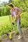 A woman in casual clothing is using a gardening tool to remove weeds in a well-maintained garden. The background is green and overgrown.