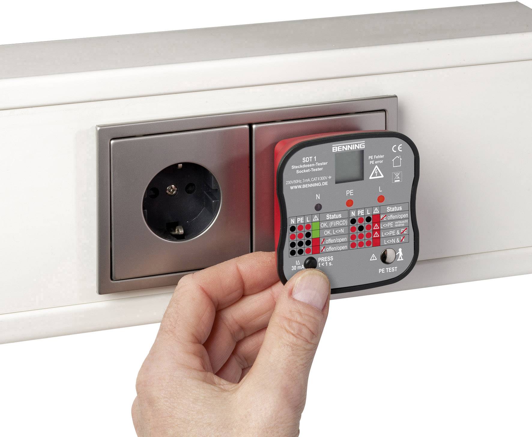 A hand holds a testing device against a wall socket. The device displays electrical safety results through coloured LEDs.